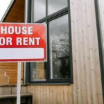 Red 'House for Rent' sign outside modern wooden house.